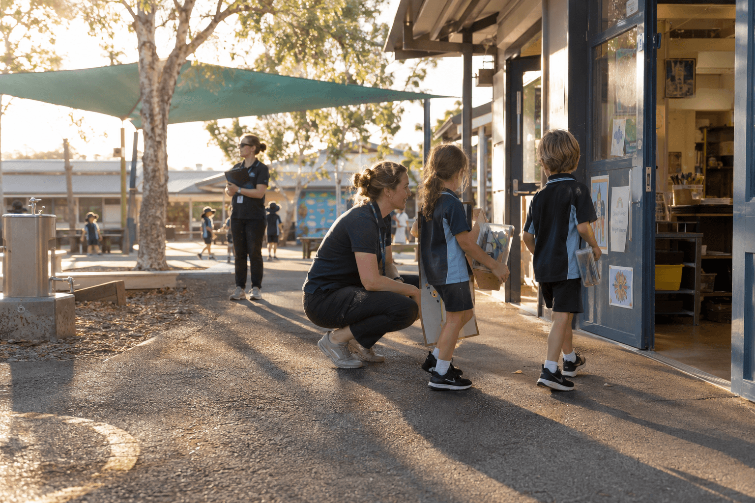 An educator crouching to chat with two children carrying art supplies into a classroom